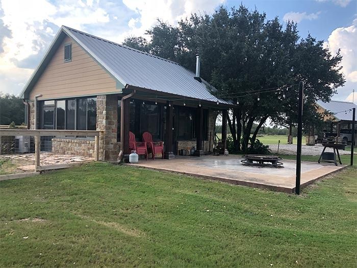 A tan cottage with a stone base and metal roof features a covered patio with red chairs, facing a lawn and large tree.