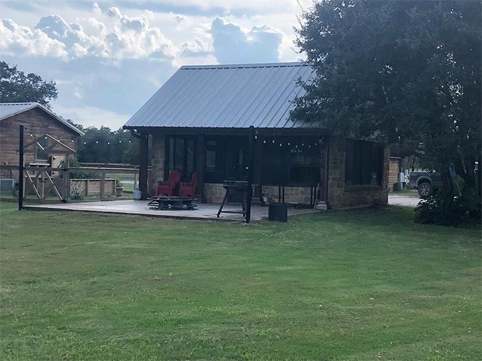 A small stone cabin with a metal roof and a paved patio featuring outdoor chairs and string lights on a grassy lawn.