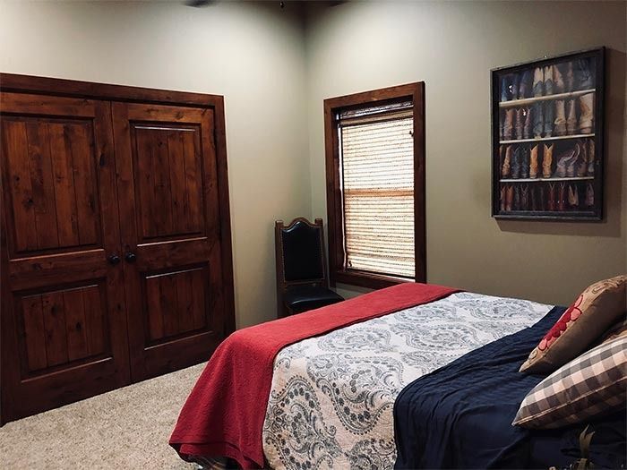 Bedroom with dark wood doors, a window with bamboo shades, a bed with red and patterned bedding, and wall art of boots.