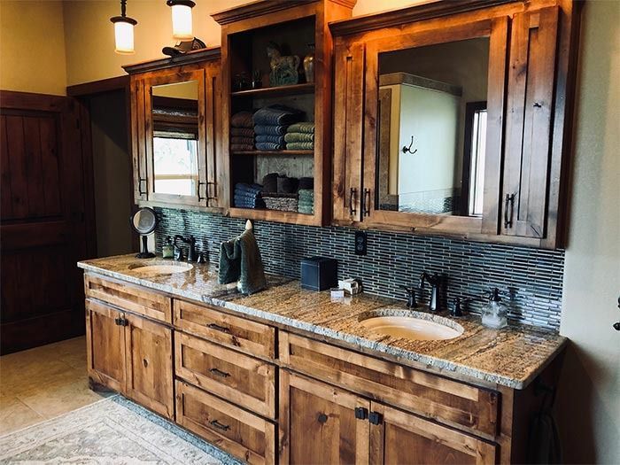 A rustic bathroom vanity with wooden cabinets, stone countertops, two sinks, and a dark mosaic tile backsplash.