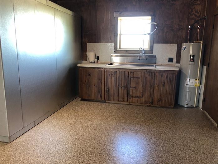 A small kitchen with wood-paneled walls, metal cabinets, a sink, and a large water heater on a speckled floor.