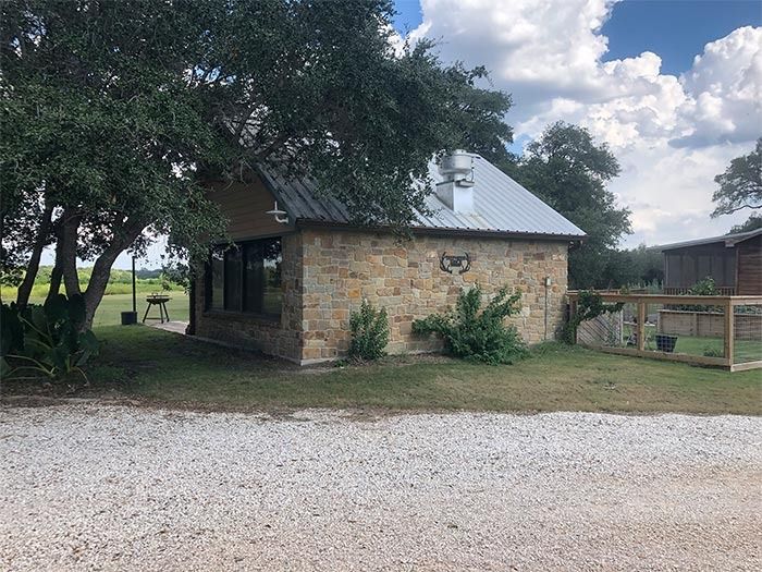 A stone building with a metal roof surrounded by trees and a gravel driveway on a sunny day.