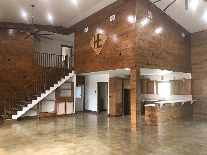 A spacious room with wood-paneled walls, a staircase leading to a loft, concrete floors, and a kitchen area with a bar.