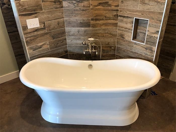 A white, freestanding oval bathtub sits in front of a curved wall tiled with brown, wood-look planks.