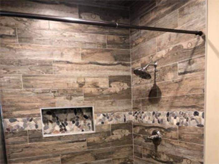 A shower stall with brown, wood-look tile walls, a horizontal pebble-tile accent strip, a small niche, and a shower head.
