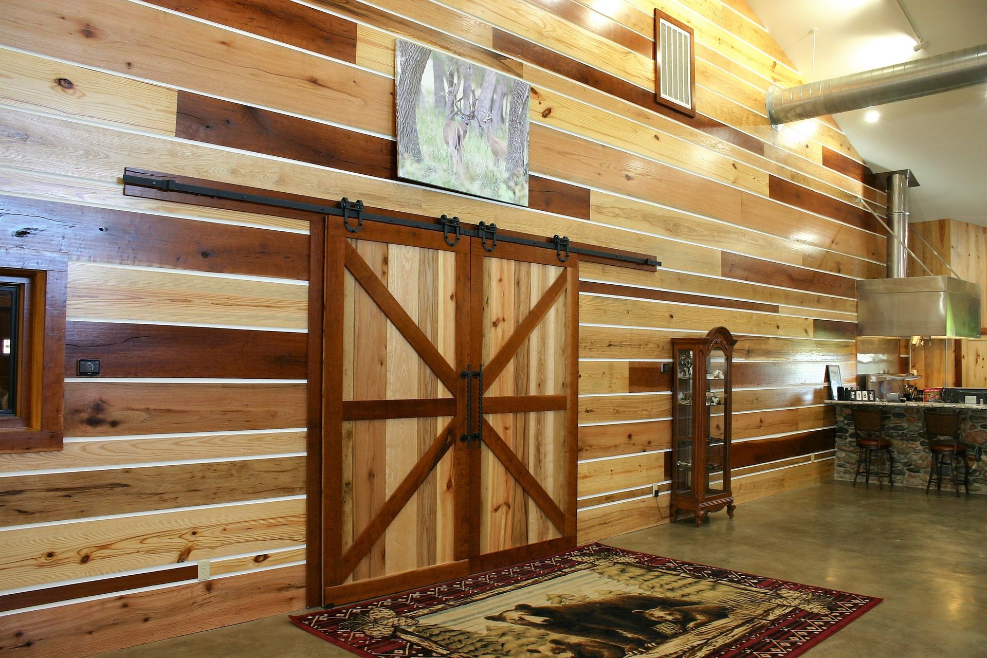 A rustic room features horizontal, multi-toned wood wall planks, a large sliding barn door, and a patterned rug on concrete.