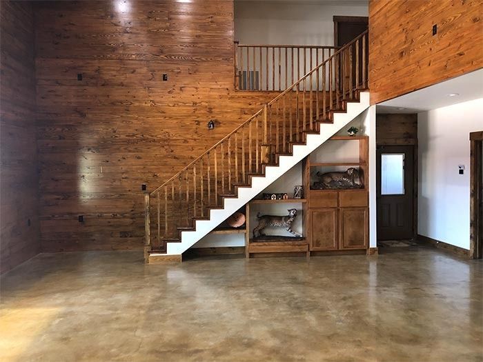 A large room with wooden walls and concrete floors, featuring a staircase with built-in wooden shelving underneath.
