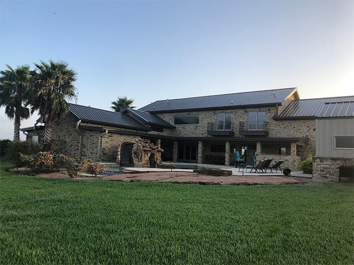 A stone two-story house with a metal roof and an outdoor patio, surrounded by green grass and palm trees.