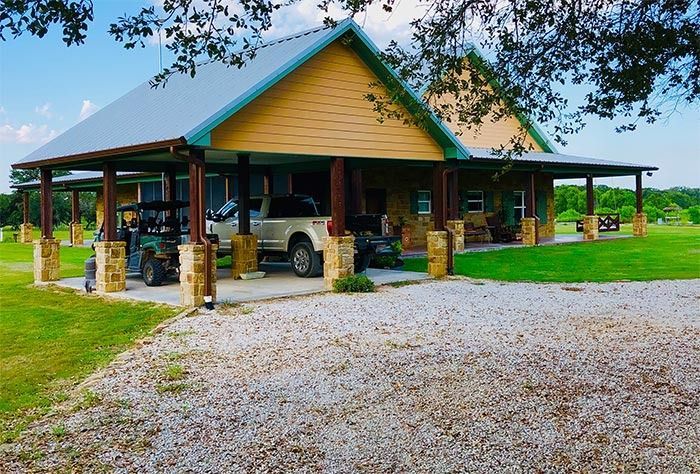 A tan house with a metal roof and a spacious stone-pillared carport featuring a pickup truck and golf cart on gravel.