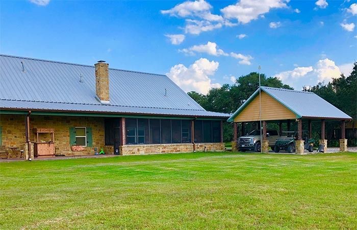 A stone house with a metal roof and a detached covered carport sits on a large green lawn under a blue, cloudy sky.