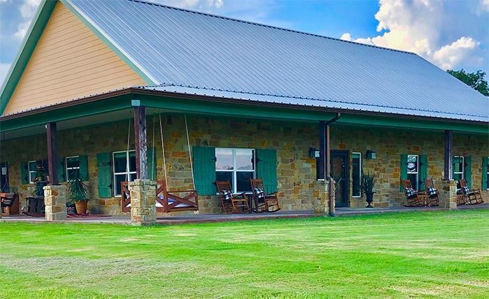 A single-story stone house with a metal roof and a spacious front porch, surrounded by a large green lawn under blue skies.