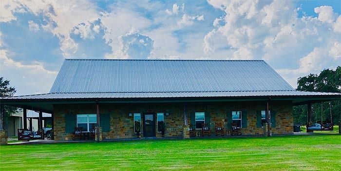A single-story stone house with a metal roof and a wide front porch sits on a grassy lawn under a cloudy blue sky.