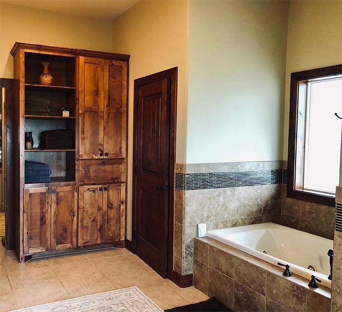 A bathroom featuring a tall, rustic wooden cabinet next to a dark wood door and a tile-surrounded bathtub near a window.