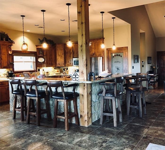 A rustic kitchen featuring wooden cabinets, a stone island with bar stools, pendant lighting, and dark tiled flooring.