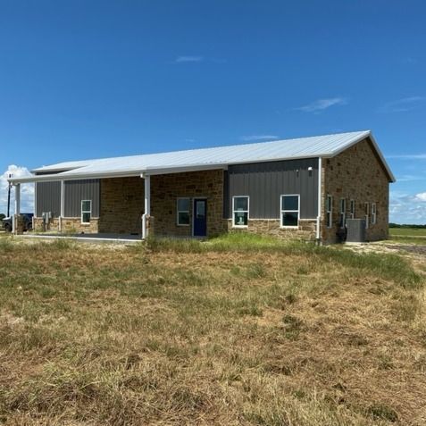 A modern building with stone and dark gray metal siding under a clear blue sky, surrounded by a dry grass field.