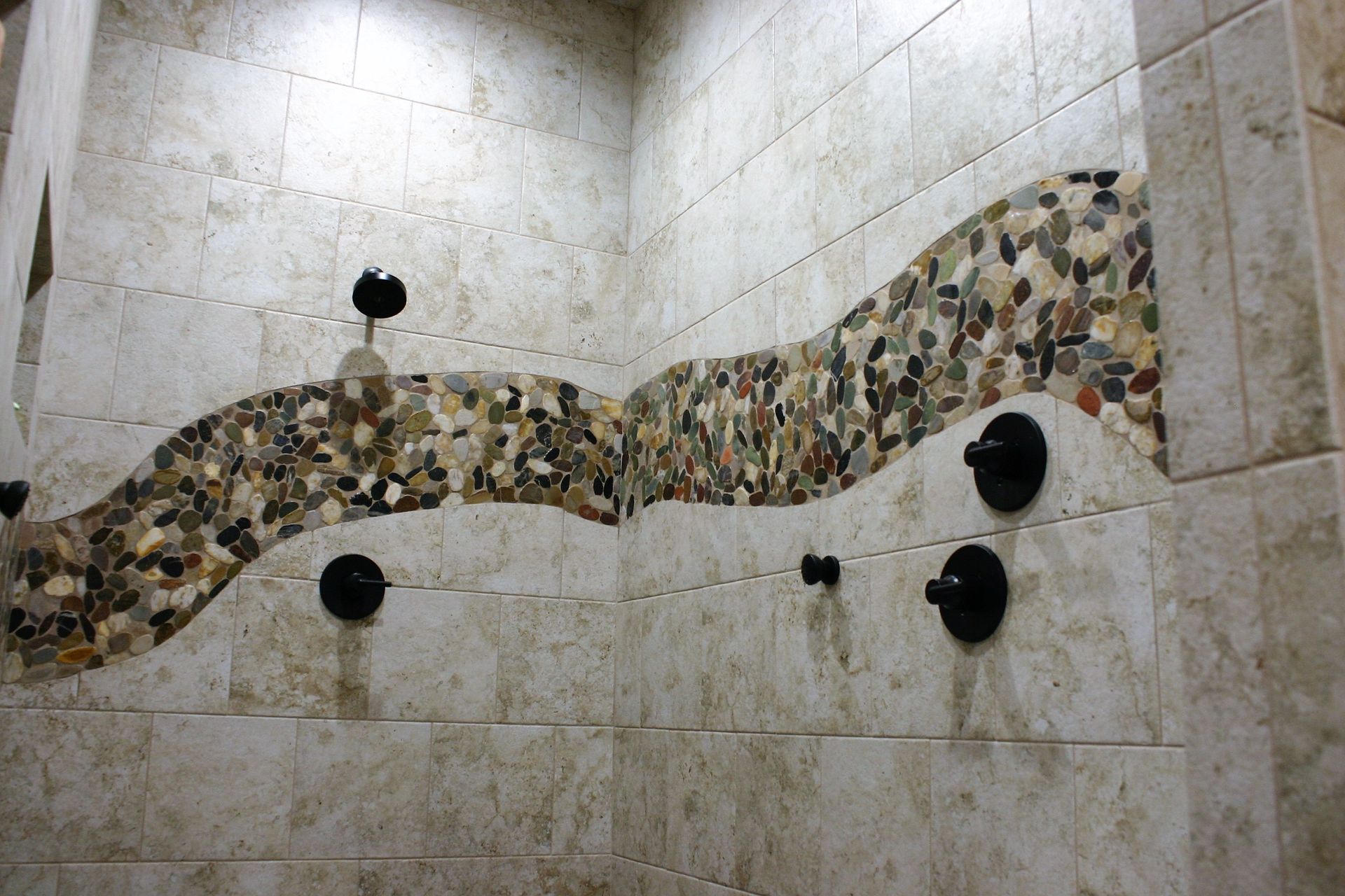A shower wall tiled in light beige stone with a wavy, decorative border of multicolored river stones and black fixtures.