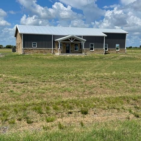 A modern, single-story metal building with stone siding and a metal roof, centered on a large, grassy rural lot.