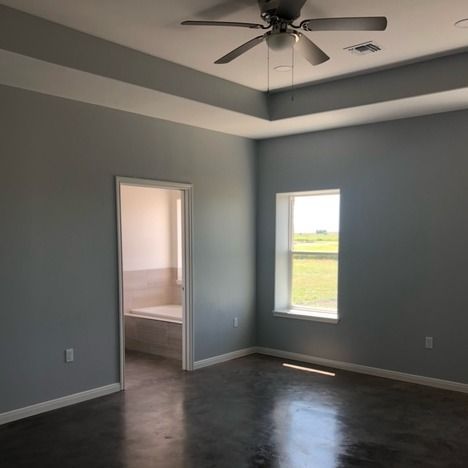 A room with light grey walls, dark concrete floors, a tray ceiling with a fan, a window, and a doorway to a bathroom.