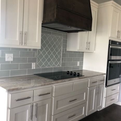 A modern kitchen with white cabinets, a large dark wood range hood, grey subway tile backsplash, and stainless oven.