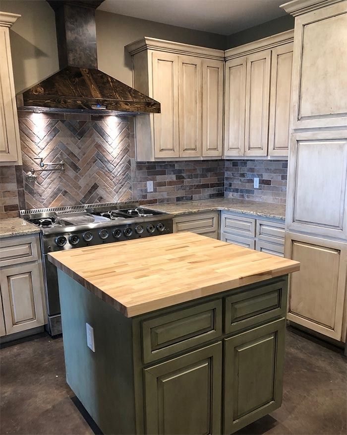 A kitchen with distressed white cabinets, a green island with a butcher block top, and a brick-tiled backsplash.