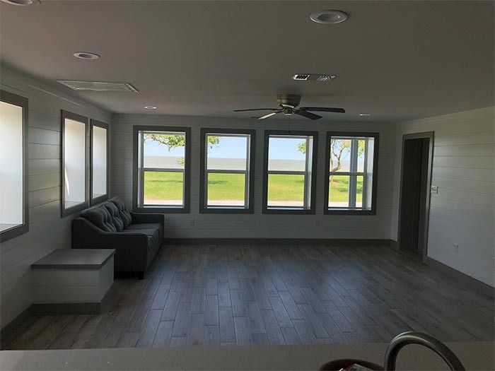 A living room with white shiplap walls, grey wood floors, a grey sofa, and four windows overlooking a grassy landscape.
