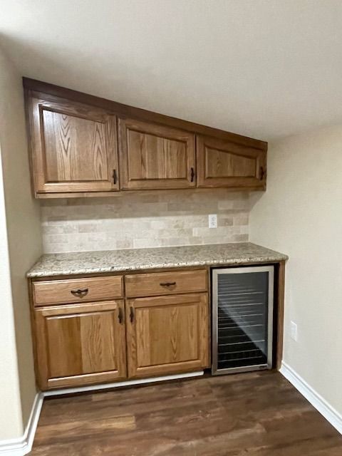 A custom home bar or kitchenette featuring wood cabinets, a granite countertop, stone backsplash, and a wine cooler.