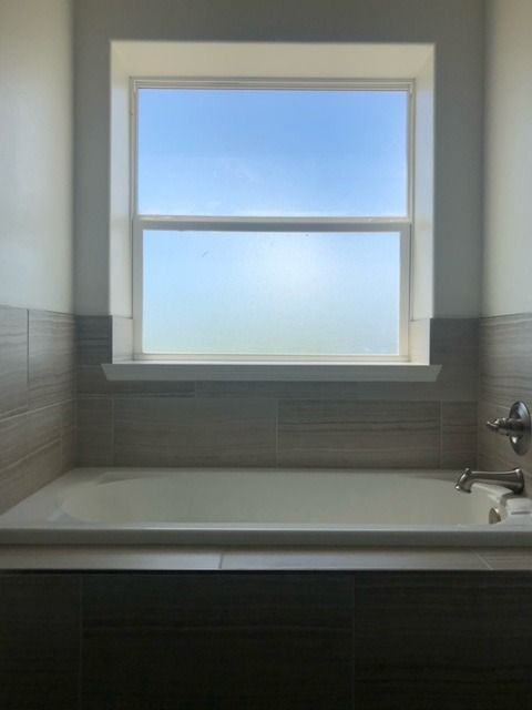 A white bathtub sits in a tiled alcove beneath a window looking out onto a clear blue sky.