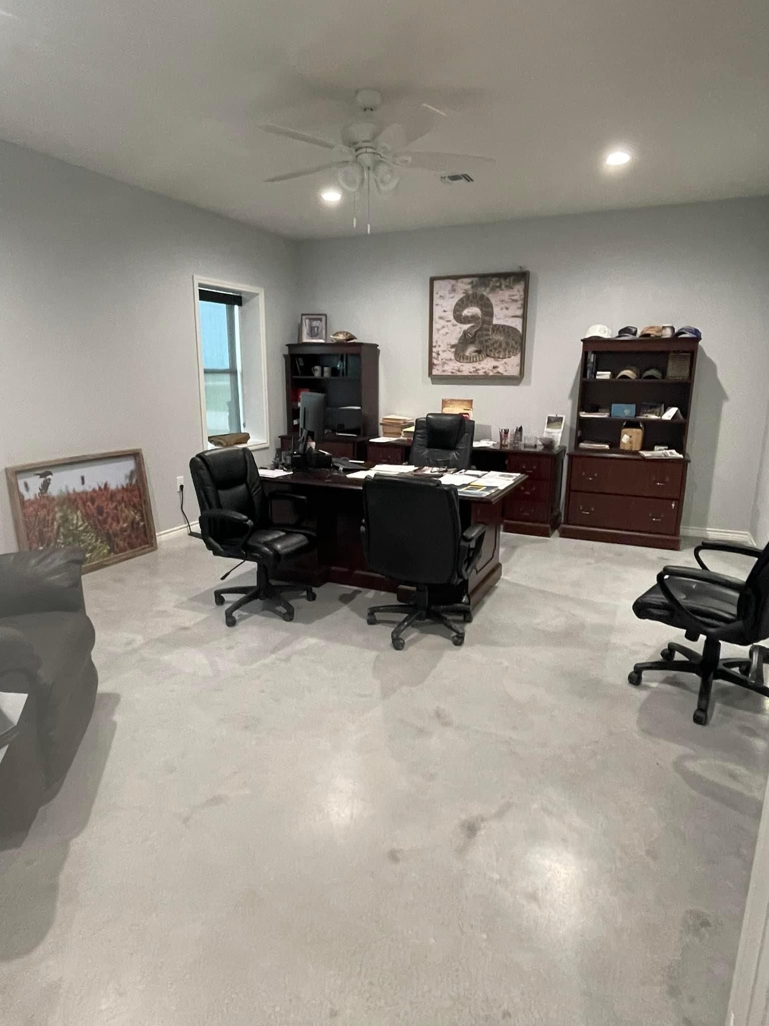 A home office with concrete floors, a desk with two black office chairs, a tall wooden bookcase, and framed wall art.