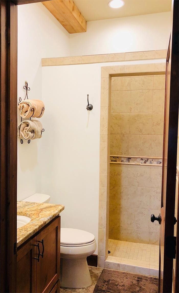A rustic bathroom with a wooden vanity, granite countertop, white toilet, and a tiled shower stall.