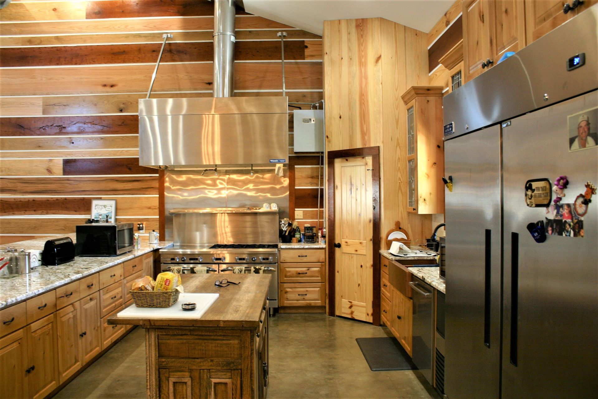 A rustic kitchen featuring wooden walls, granite countertops, stainless steel appliances, and a central wooden island.