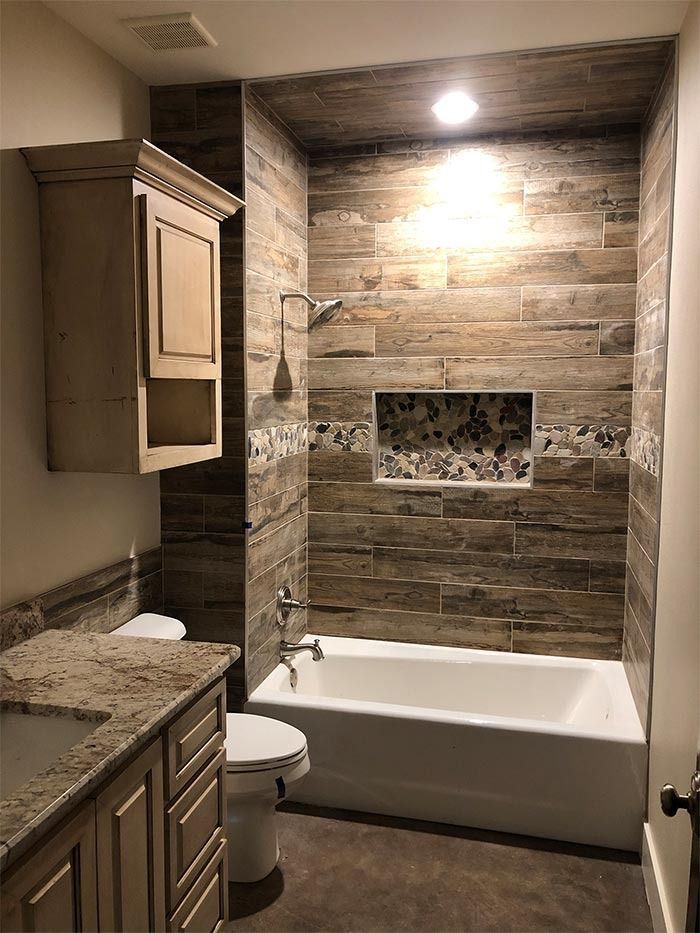 A bathroom with wood-look tiled walls in the bathtub area, featuring an inset shelf, a white tub, and a vanity cabinet.