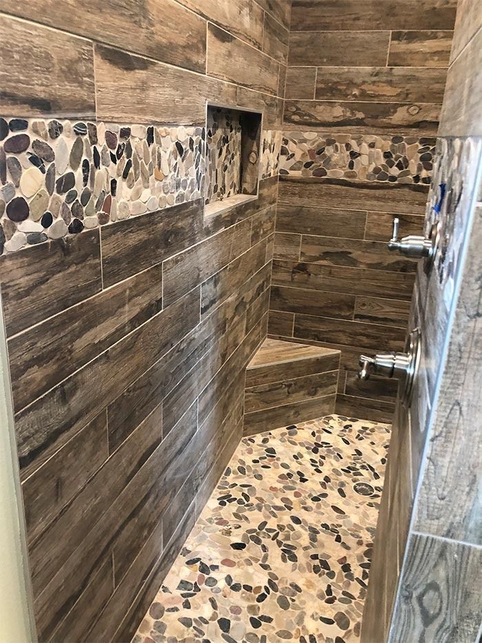A shower interior with brown wood-look tiles, a pebble-stone floor, a matching horizontal tile accent, and a corner seat.