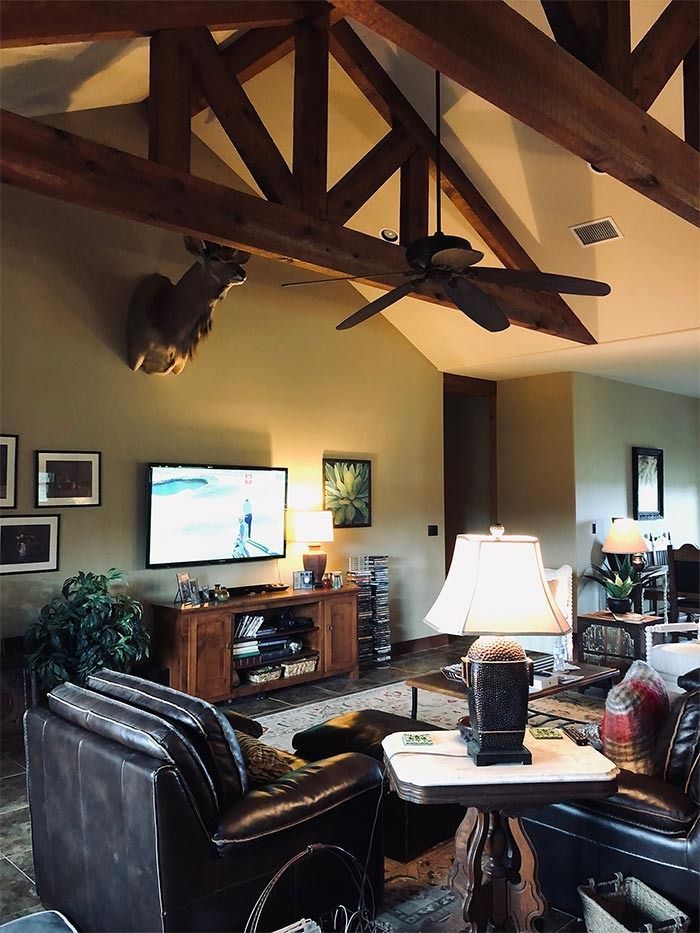 A cozy living room with exposed wood ceiling beams, a mounted deer head, a TV, leather armchairs, and a table lamp.