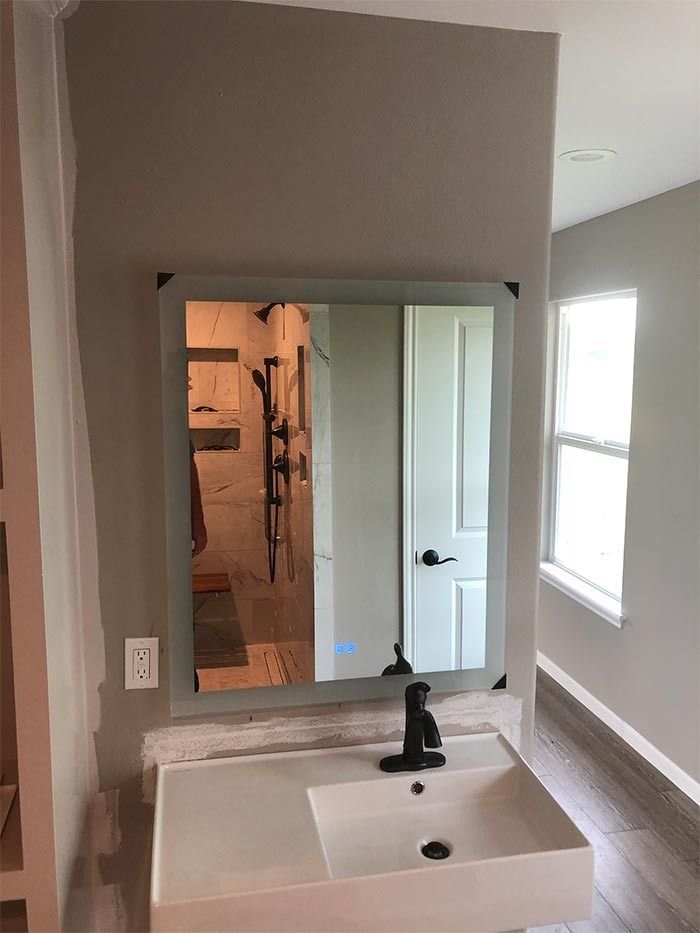 A wall-mounted bathroom sink with a black faucet sits below a rectangular LED mirror in a gray room with a window.