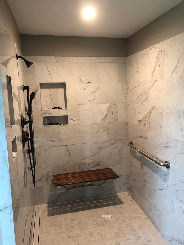 A walk-in shower with white marble-style walls, a wooden fold-down seat, grab bar, black shower fixtures, and two niches.