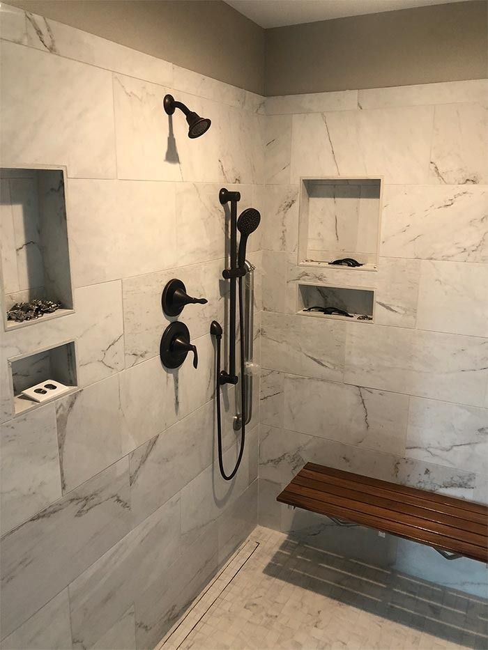 A modern marble-tiled shower featuring bronze fixtures, a handheld sprayer on a slide bar, recessed niches, and a wood bench.