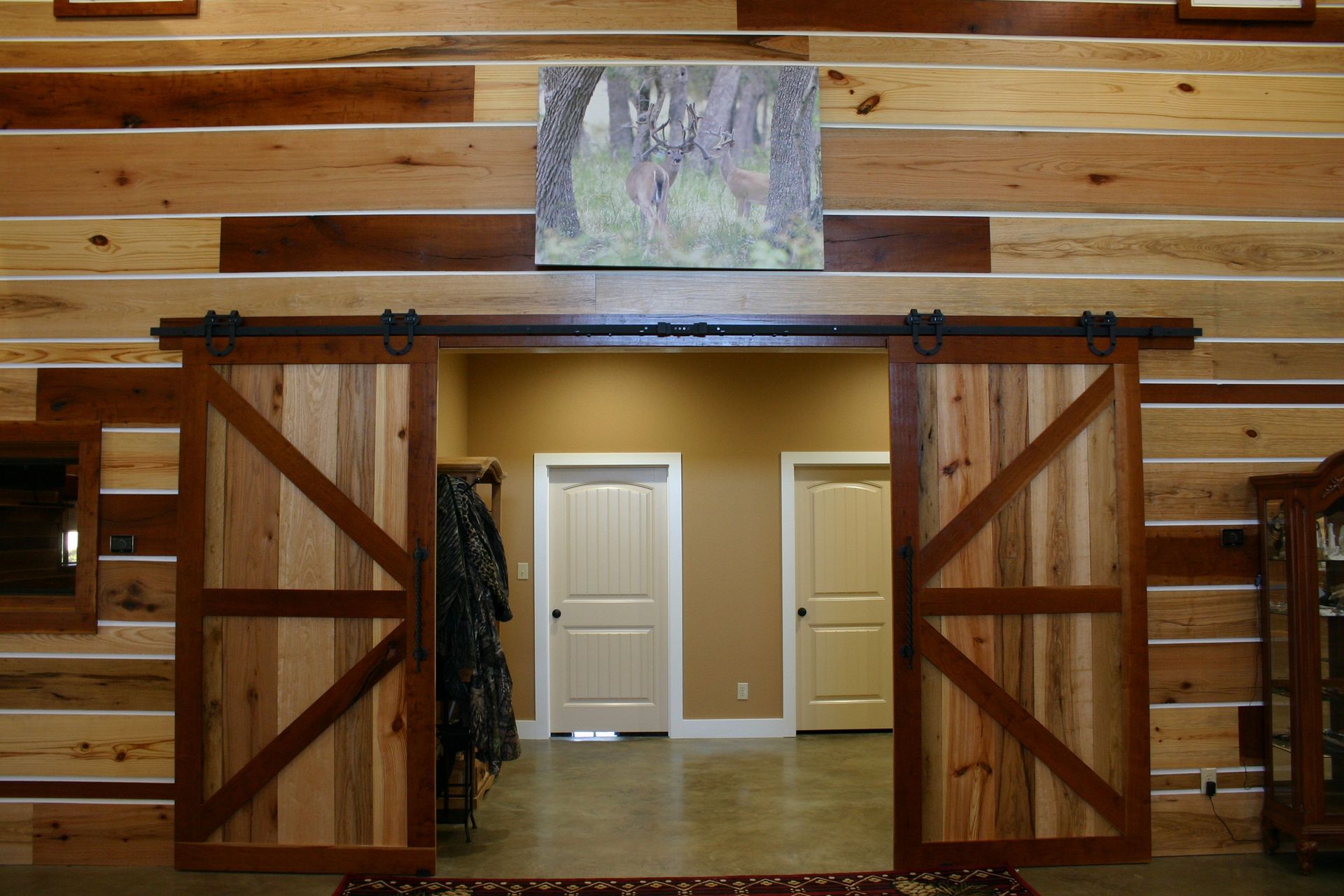 Rustic sliding barn doors open to a hallway with two white doors, set against a log-style paneled wall with a forest art.