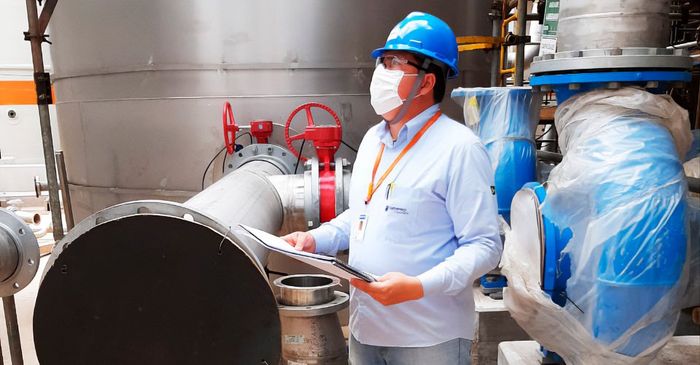Um homem usando capacete e máscara está em uma fábrica olhando para uma prancheta.