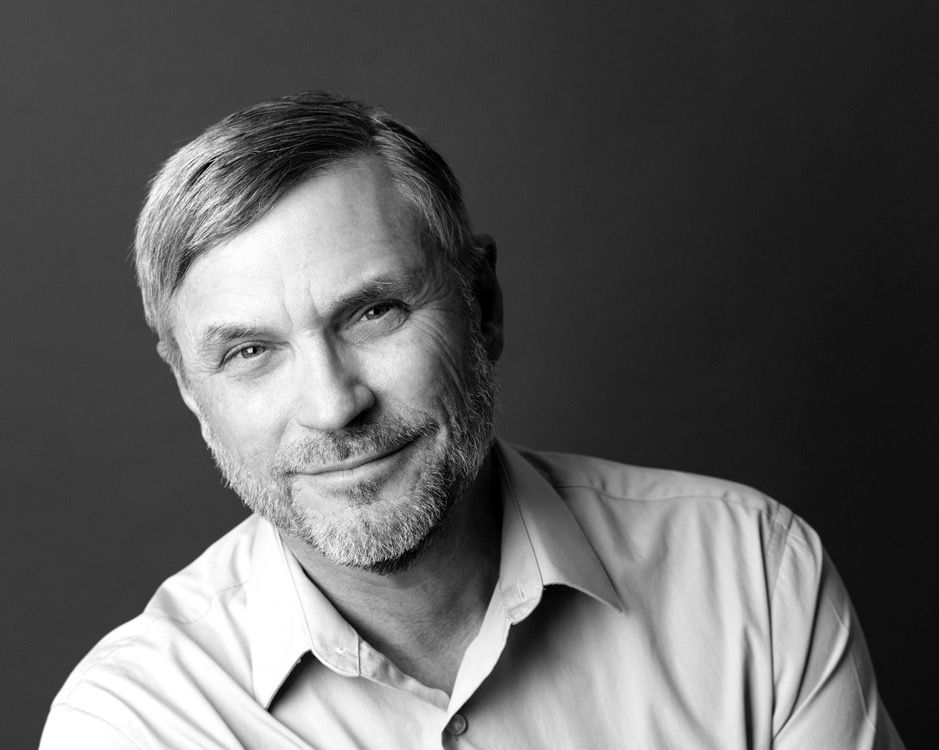 Smiling man with gray hair and beard, wearing a light-colored button-up shirt, against a dark background.
