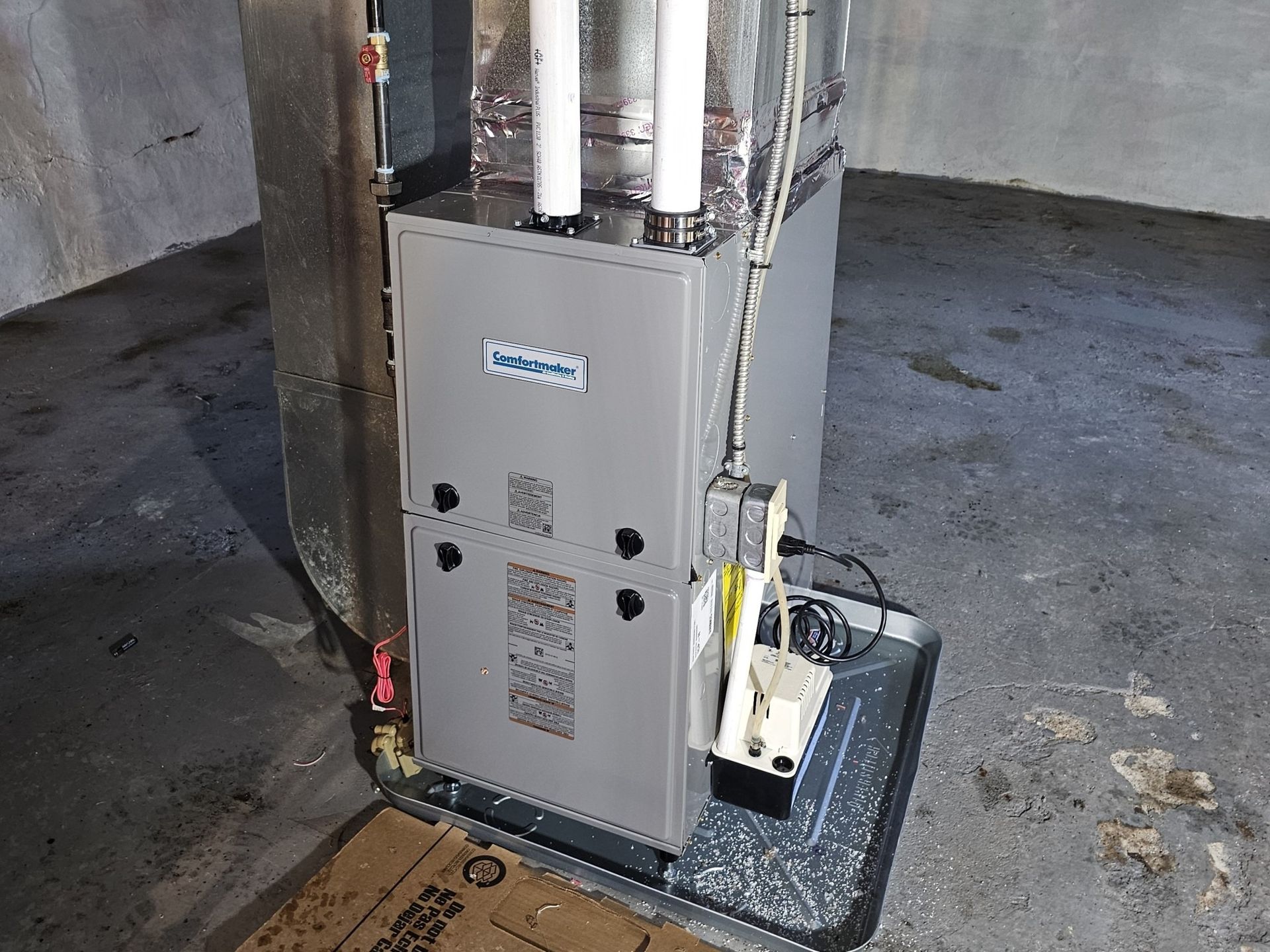 A gray furnace is sitting in a basement next to a cardboard box.