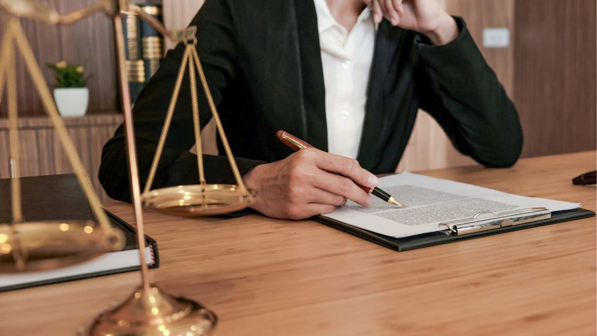 Lawyer in a black suit reviews documents at a wooden desk, scales of justice on the left.