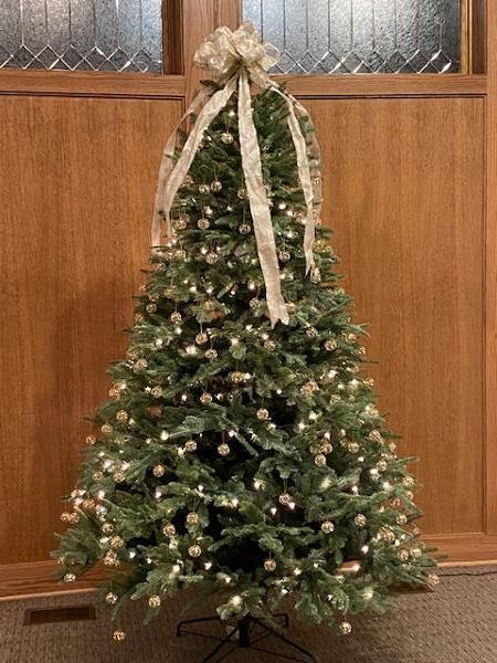 Christmas tree with white lights and bow, in front of a wood paneled wall.