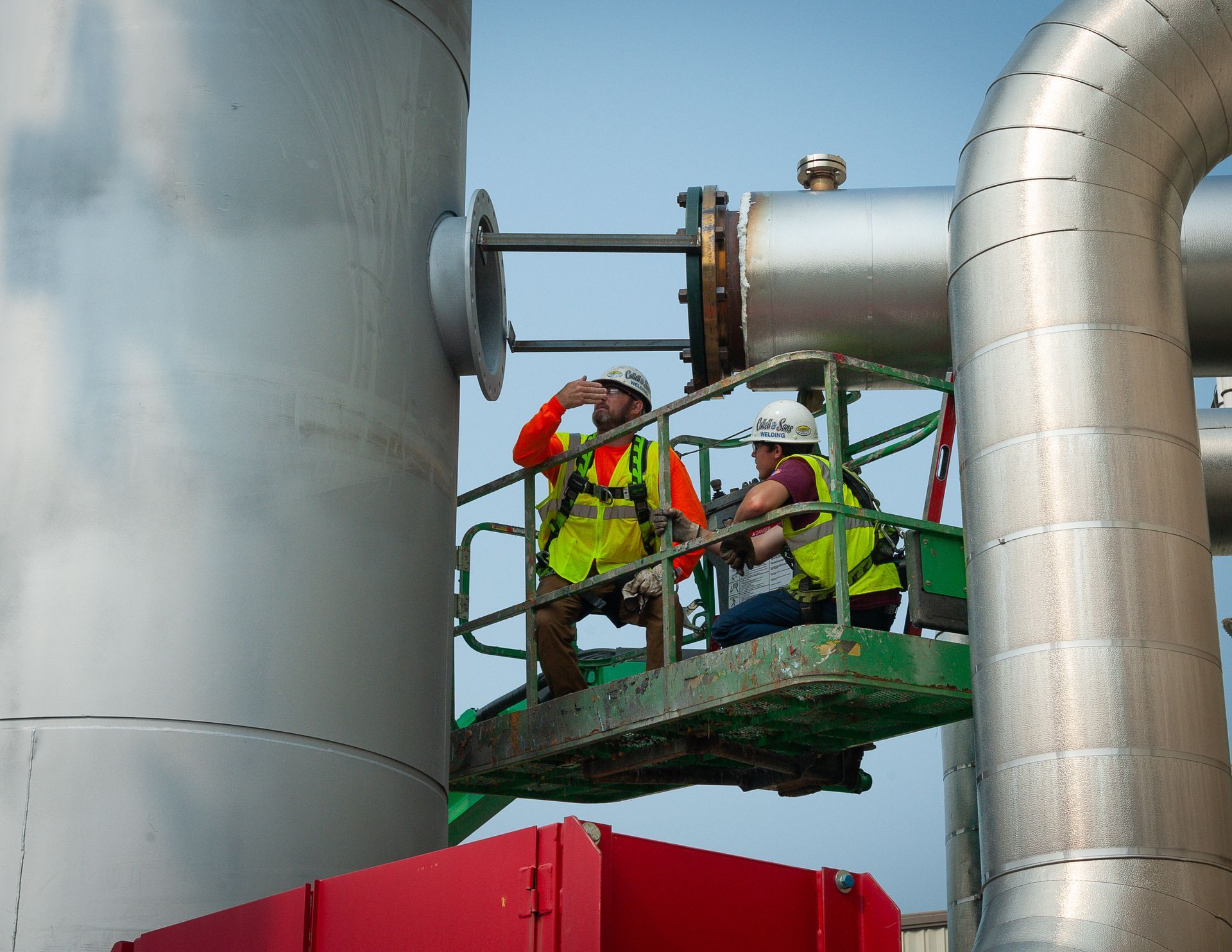 Two construction workers are standing on a platform looking at pipes
