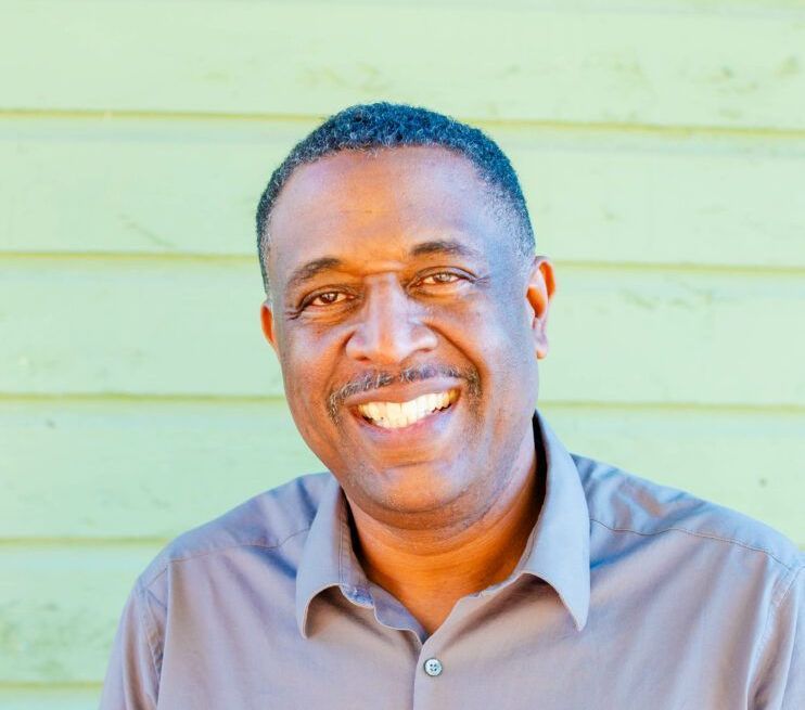 A man in a grey shirt is smiling in front of a green wall.