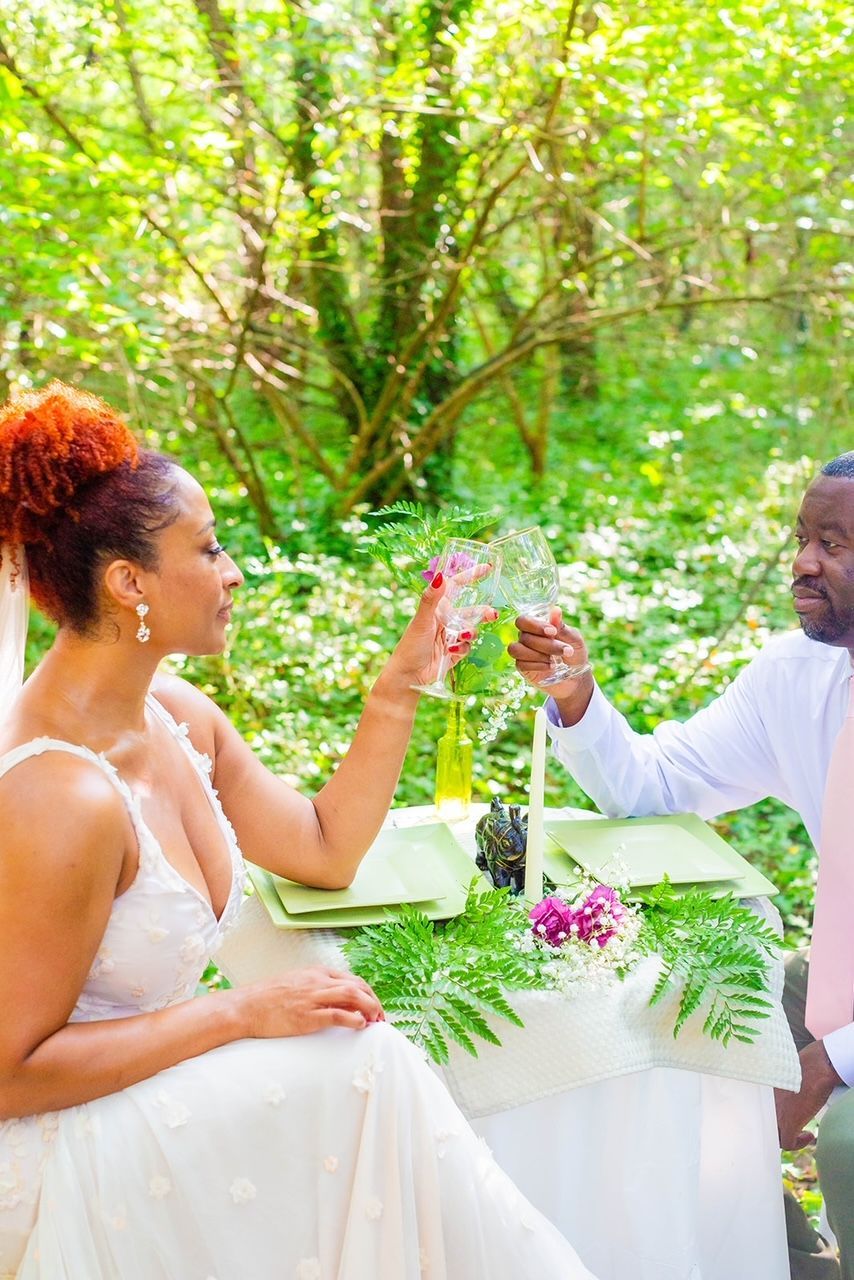 a bride and groom are sitting at a table in the woods toasting with wine glasses .