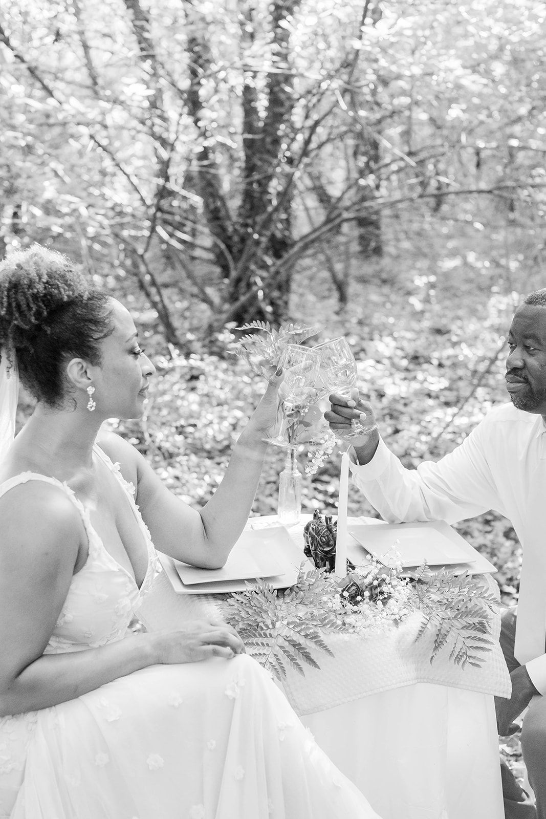 a bride and groom are sitting at a table in the woods drinking wine .