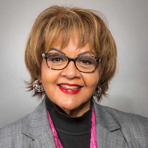 Woman in glasses smiles, wearing a gray blazer, black top, and pink scarf.