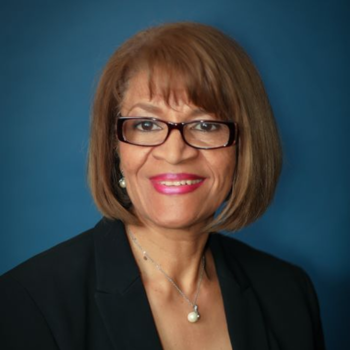 Berta Talbot in glasses, wearing a black blazer, smiling at the camera, with a blue background.
