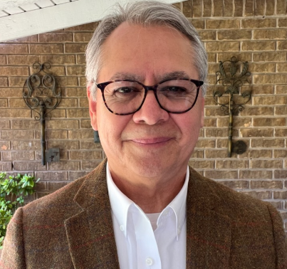 Fidencio Flores in glasses and brown jacket smiles in front of a brick wall with decorative metal keys.