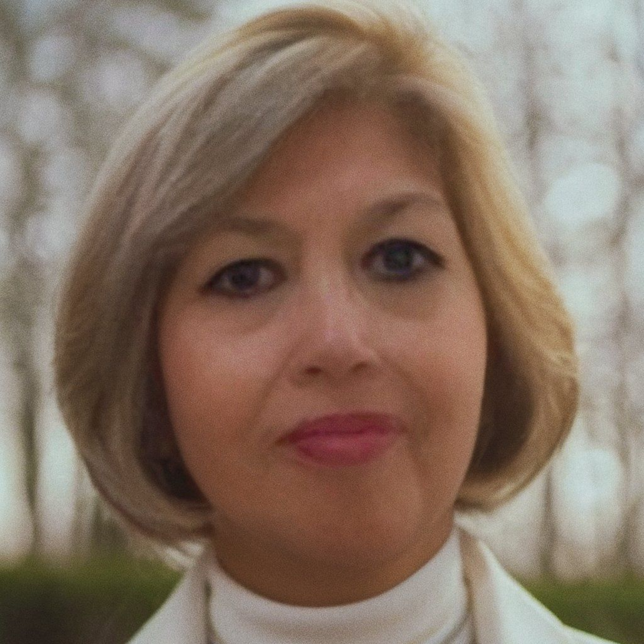 Sarah Vasquez with blonde, chin-length hair, smiling slightly, in a white turtleneck, outdoors.
